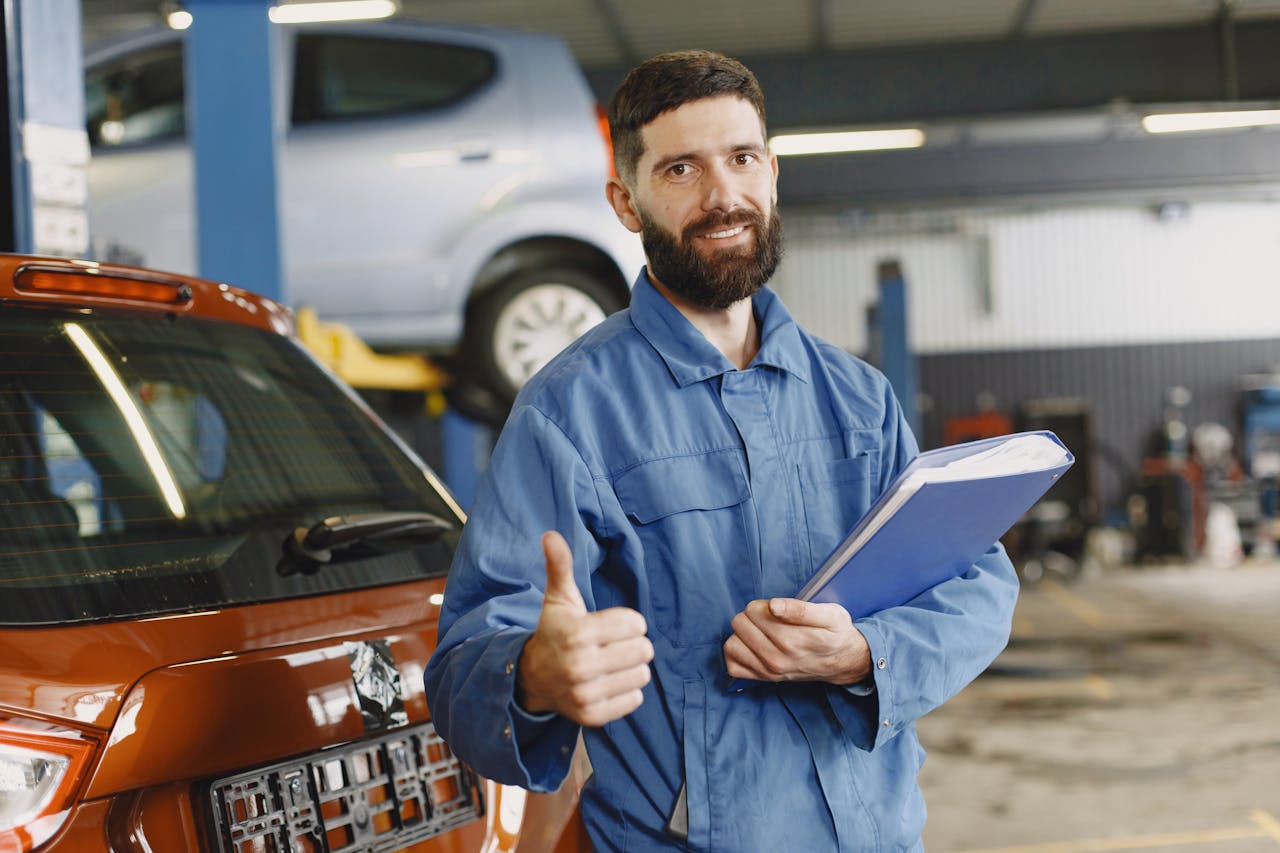 Auto mechanic in blue coveralls with clipboard giving thumbs up in garage.